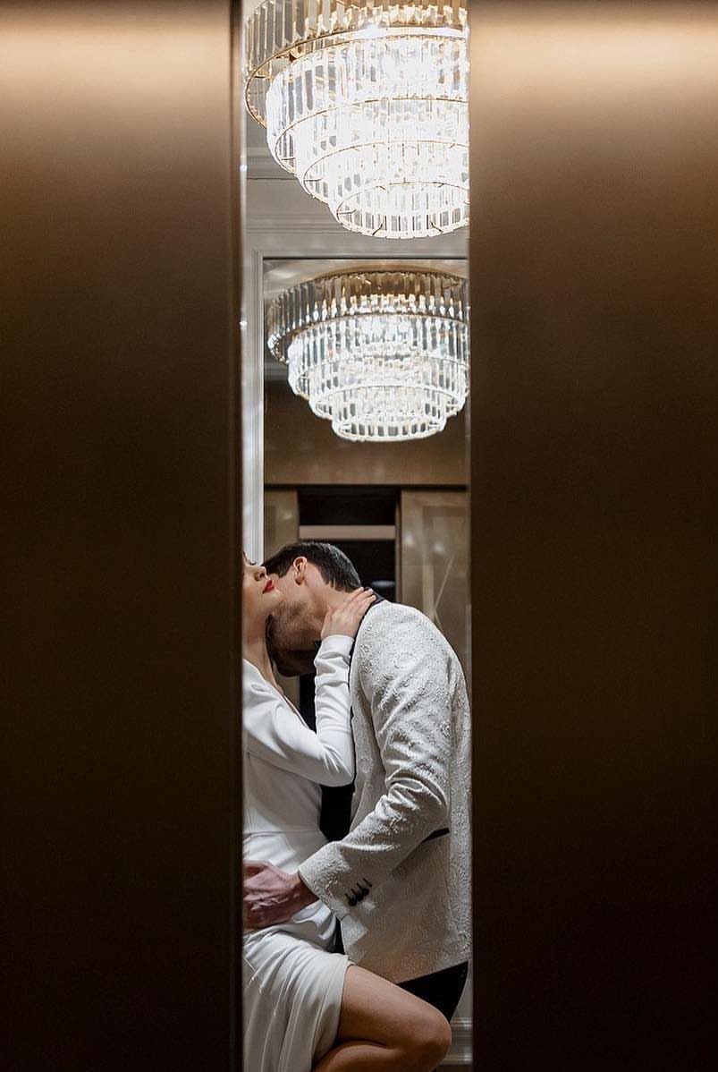 17 Couple Pictures for Your Wedding That Feel Real, Not Posed 12 A couple kissing inside an elevator viewed through a narrow door opening with a crystal chandelier.