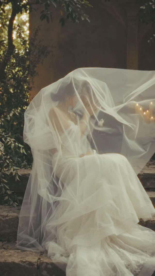 17 Couple Pictures for Your Wedding That Feel Real, Not Posed 5 A bride and groom sitting together under a large, flowing white wedding veil outdoors.