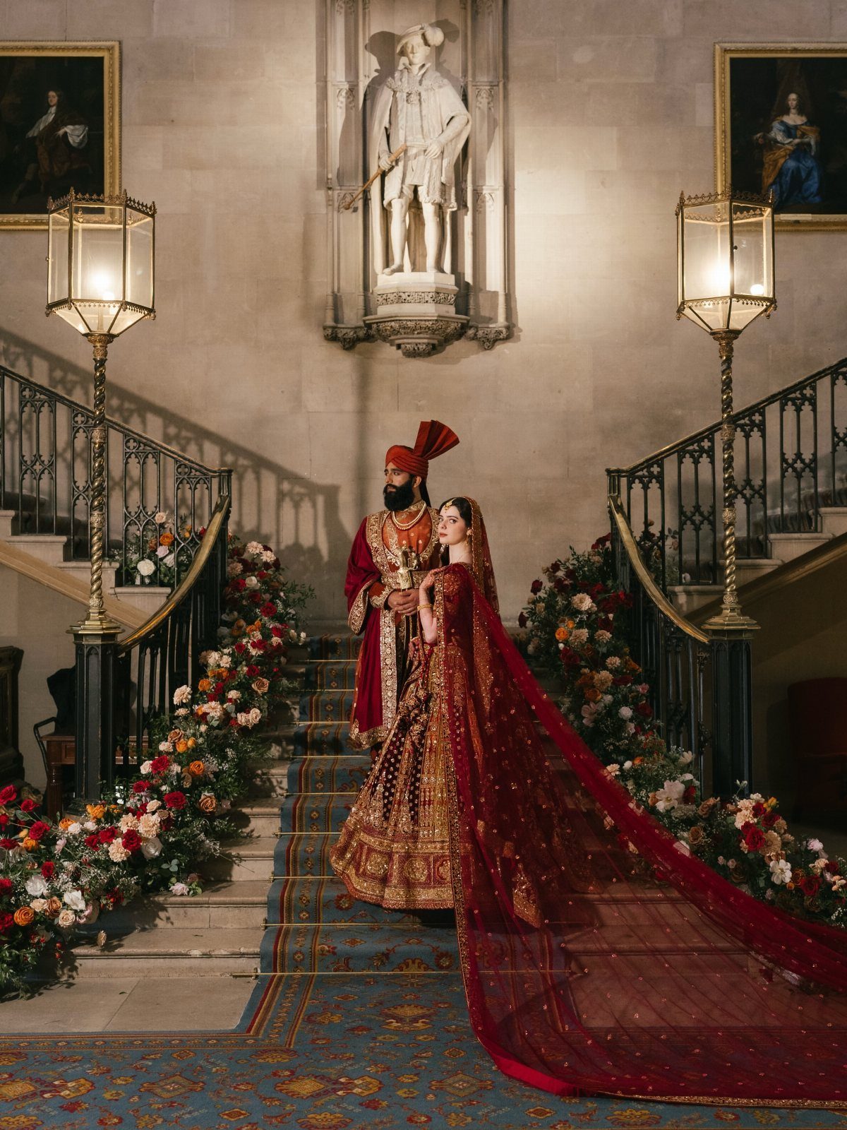 A couple in ornate red and gold wedding attire posing on a flower-decorated grand staircase.