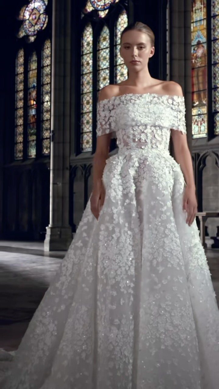 A woman wearing an off-the-shoulder white wedding gown with 3-D floral appliques in a cathedral setting.