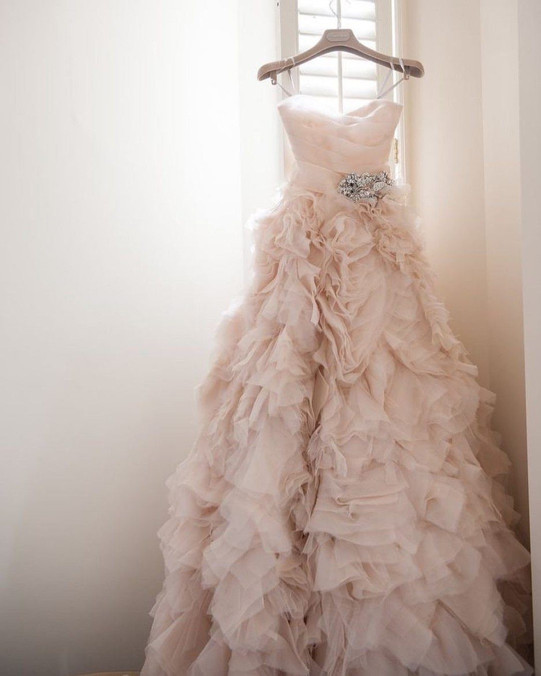 A pale pink strapless wedding gown with a voluminous ruffled skirt hanging by a window.
