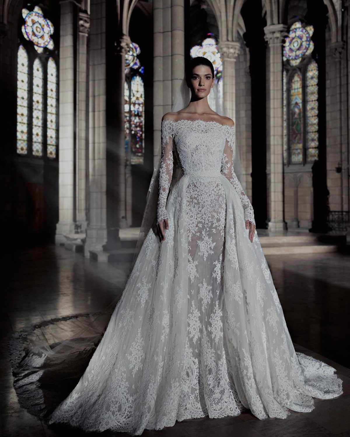 Woman in a white lace wedding gown standing in a cathedral with stained glass windows.