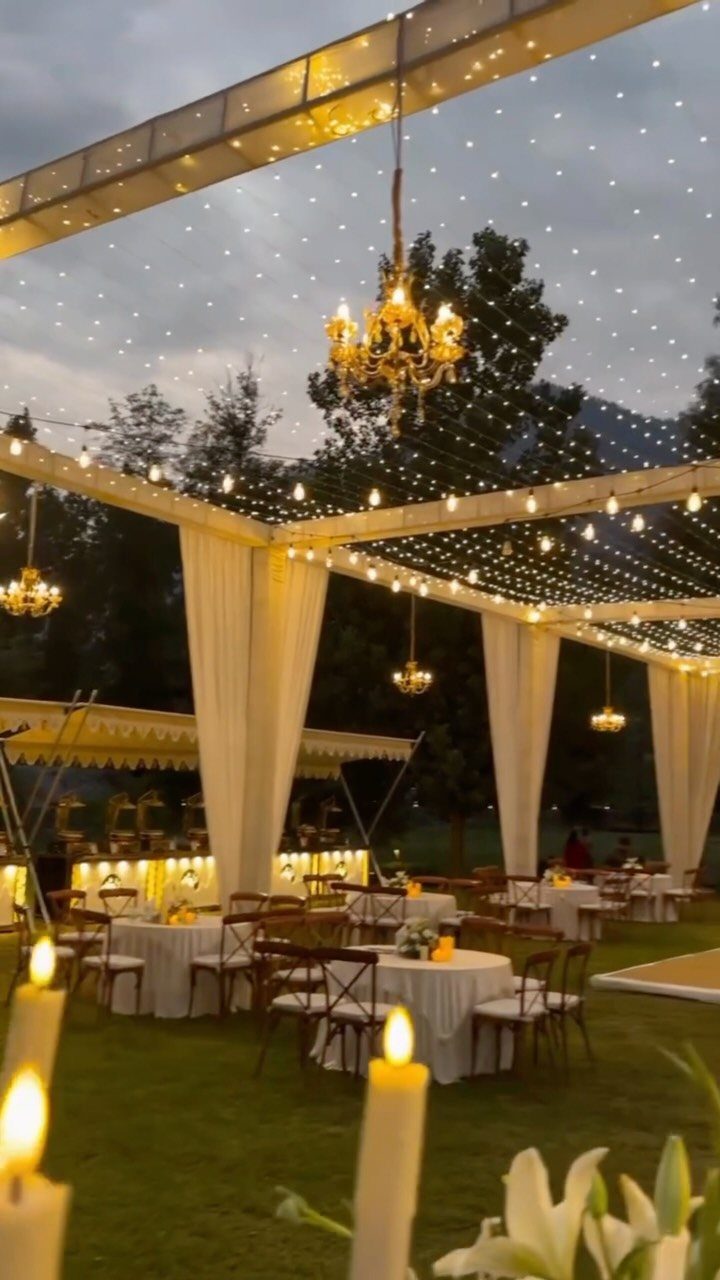 Warm fairy lights and golden chandeliers hang over white tables at an outdoor wedding reception.