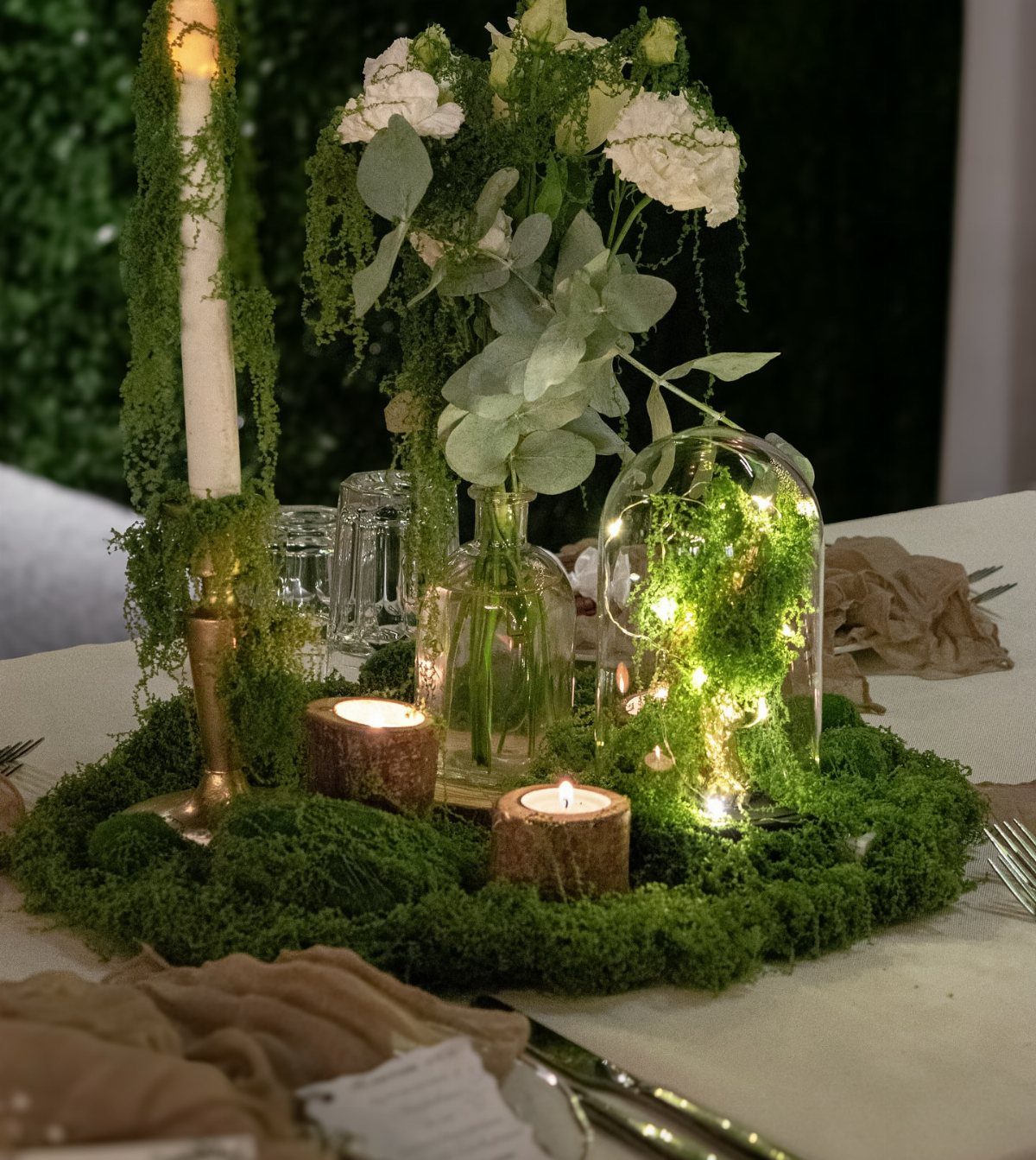 Green moss centerpiece with white flowers, candles, and fairy lights on a wedding table.
