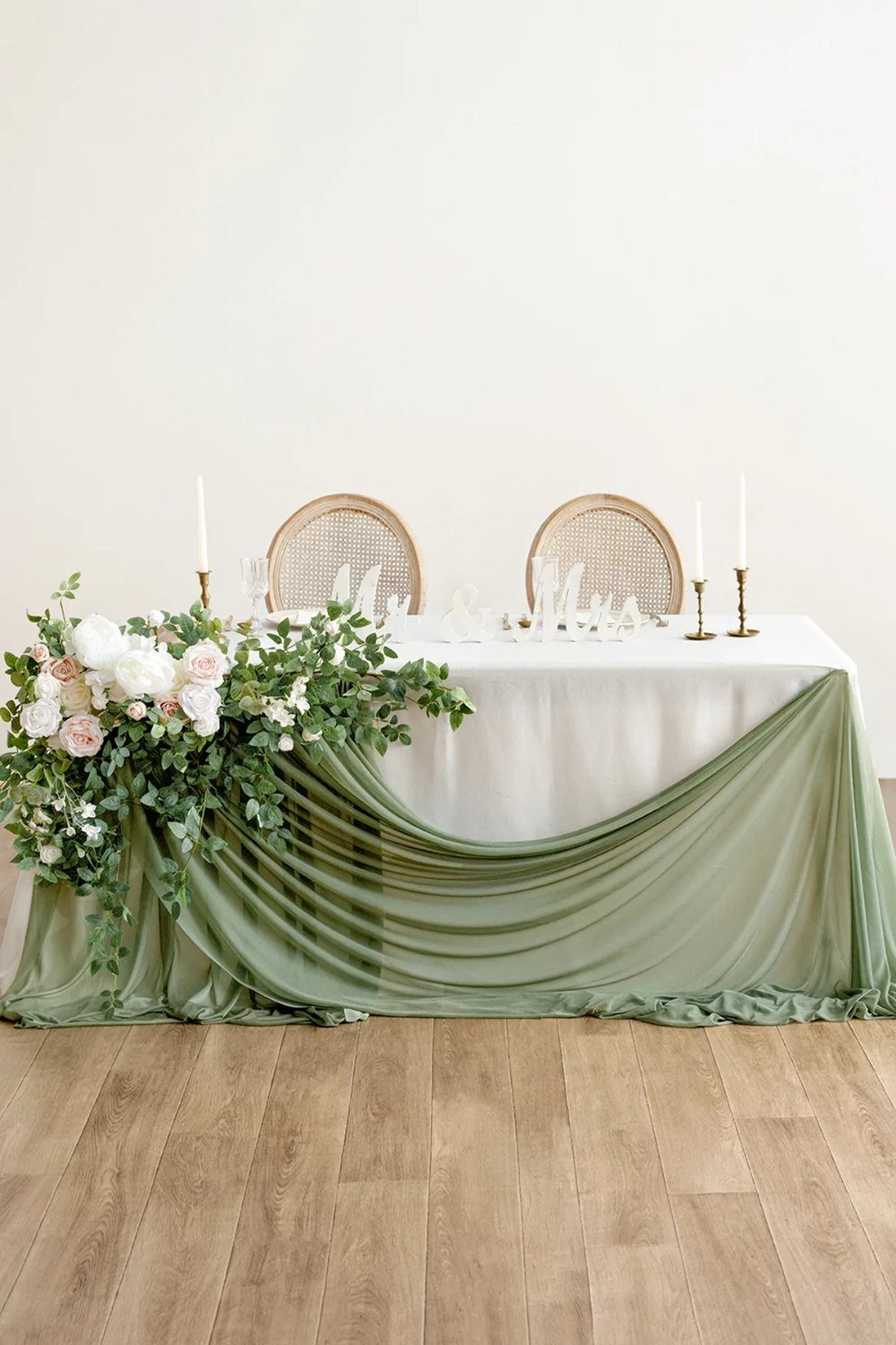 A wedding table featuring white roses, lush greenery, and sage green fabric draping.