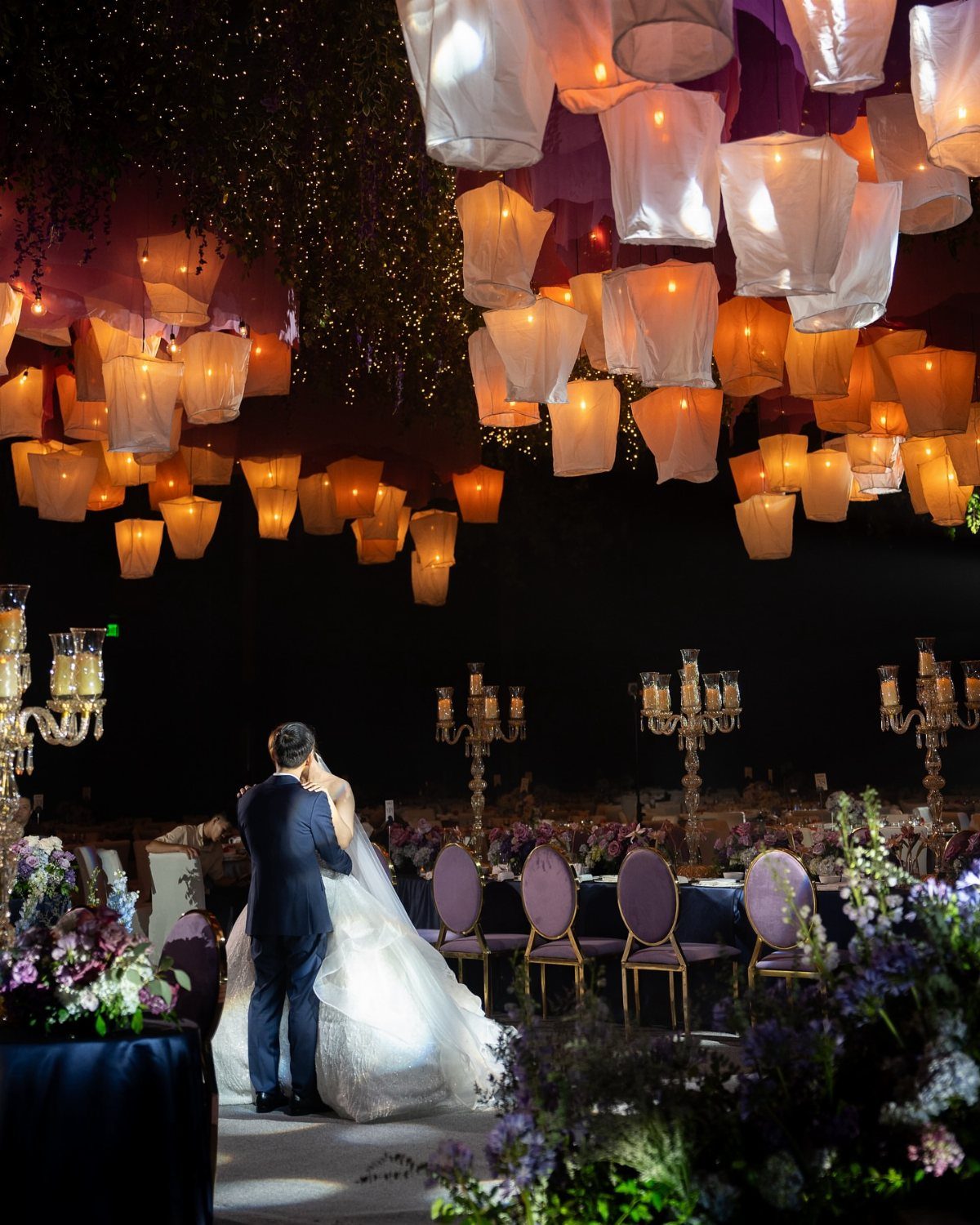 Glowing paper lanterns hang from a dark ceiling over a wedding reception setting.