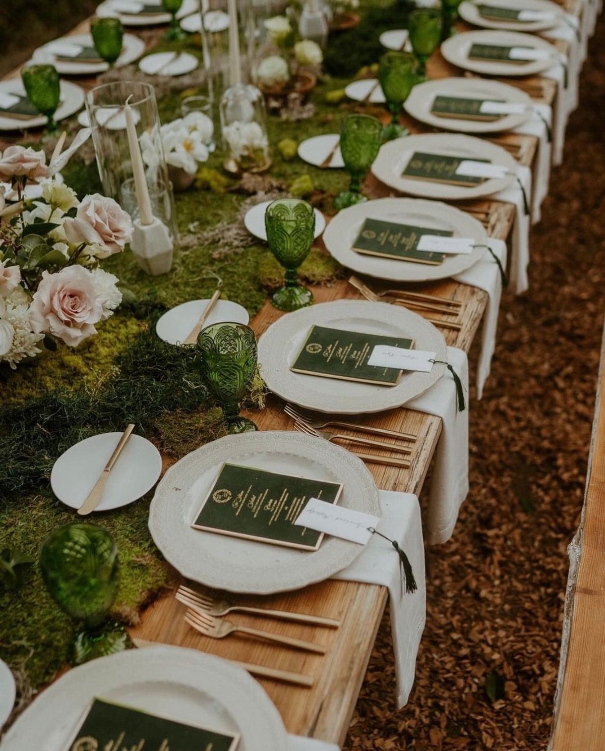 A long wooden wedding table decorated with moss, white roses, and green glassware for an outdoor event.