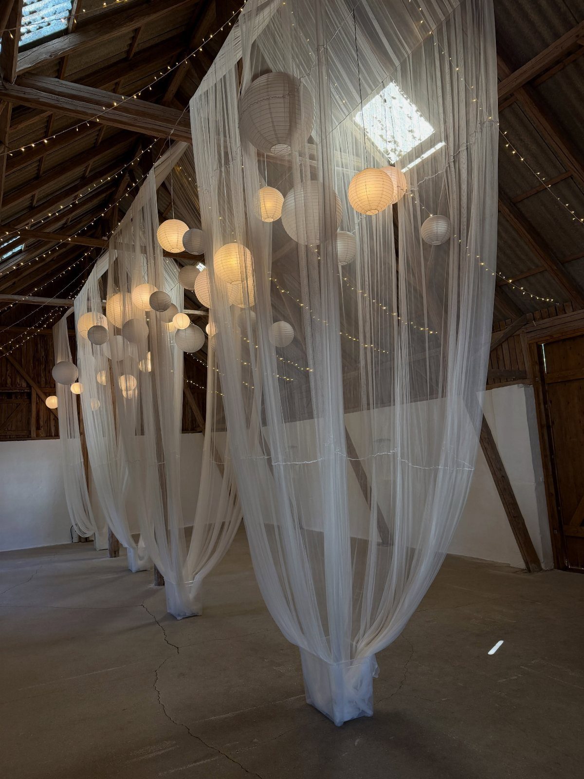 White paper lanterns hanging within draped white tulle fabric in a rustic wooden barn.