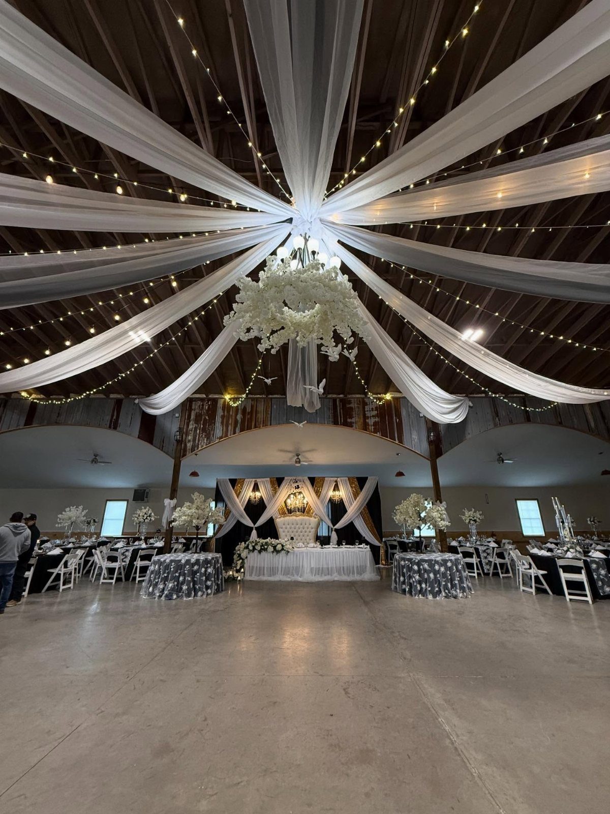 White fabric drapes and warm fairy lights radiate from a central floral chandelier in a large hall.
