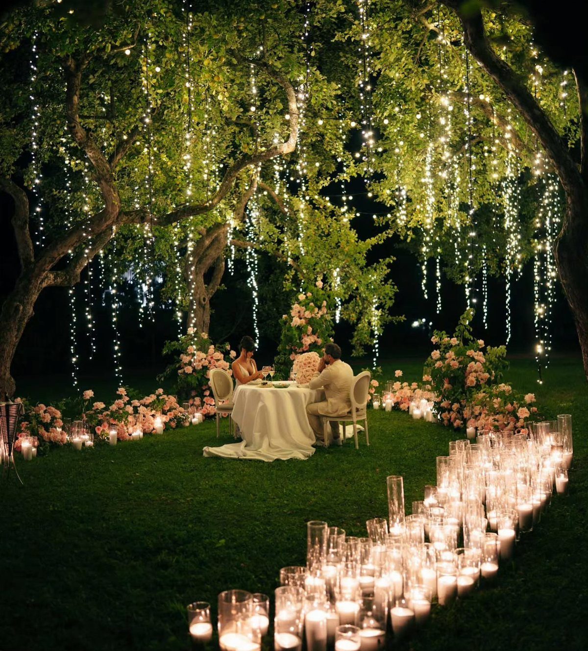 11 Forest Theme Wedding Ideas for a Natural Woodland Ceremony 1 A couple dining under a large tree decorated with hanging strands of fairy lights and glowing candles.