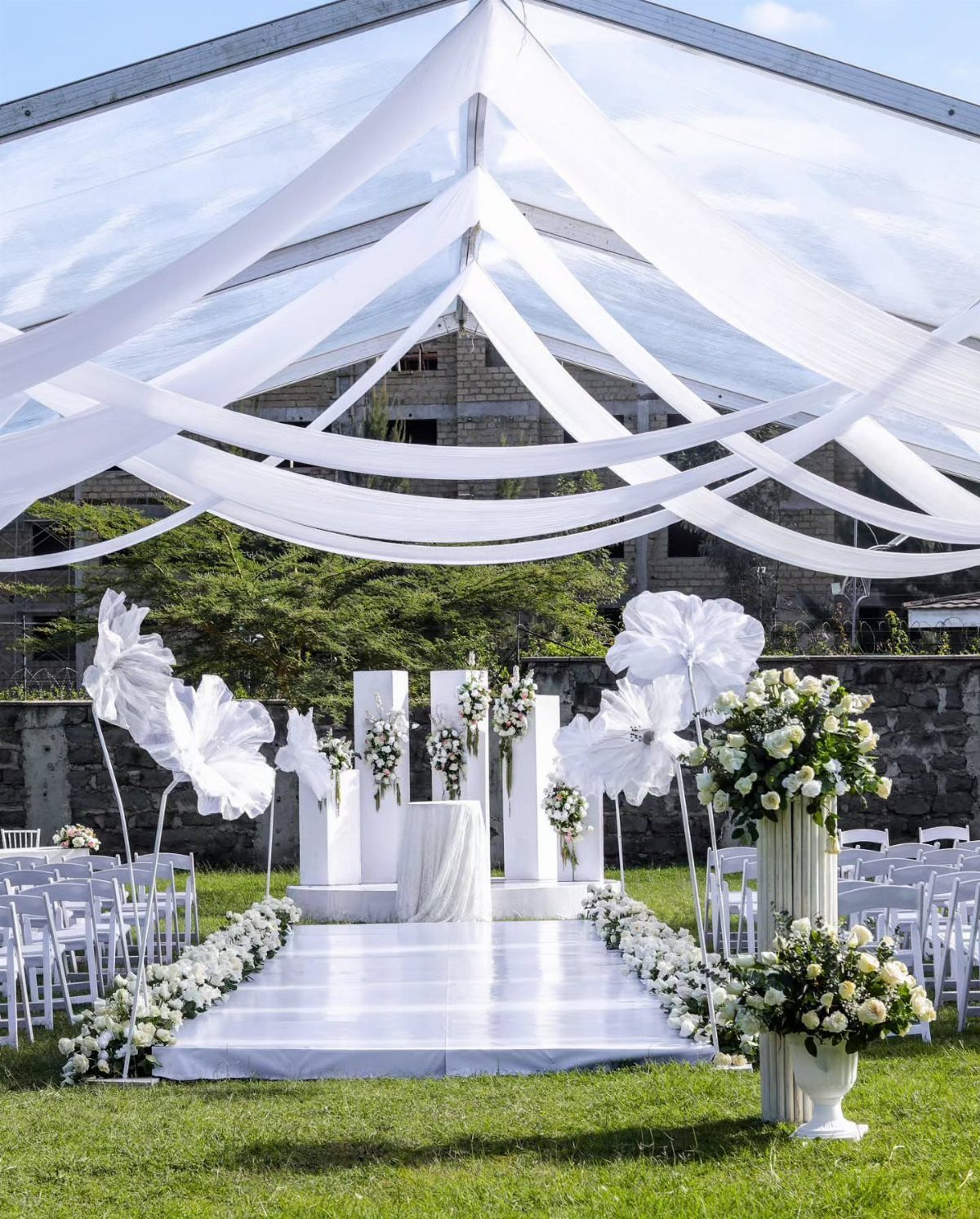 White wedding ceremony setup with a draped marquee, floral altar, and white aisle runner on green grass.