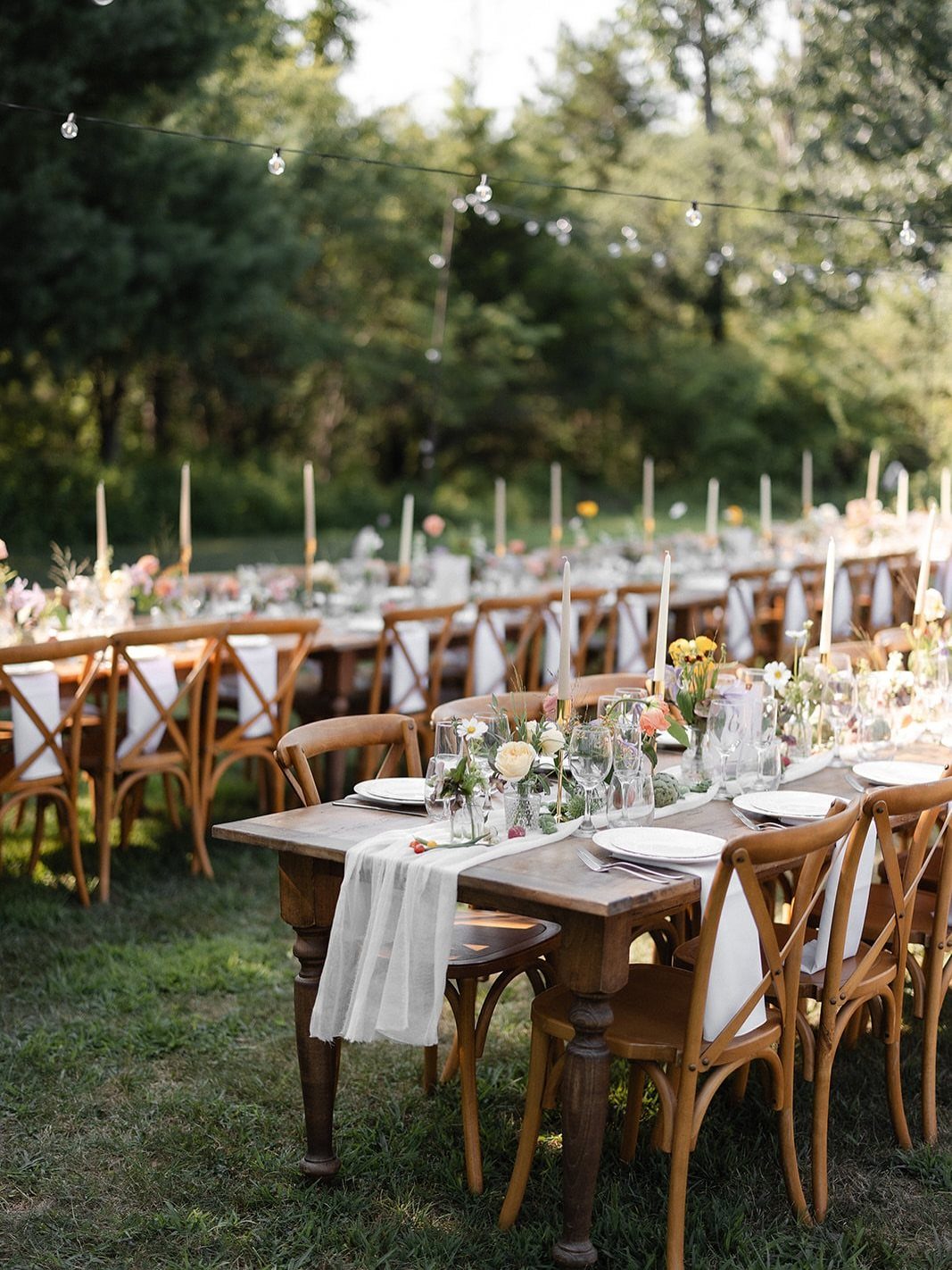 Long wooden dining tables decorated with flowers and candles set in a grassy outdoor garden area.
