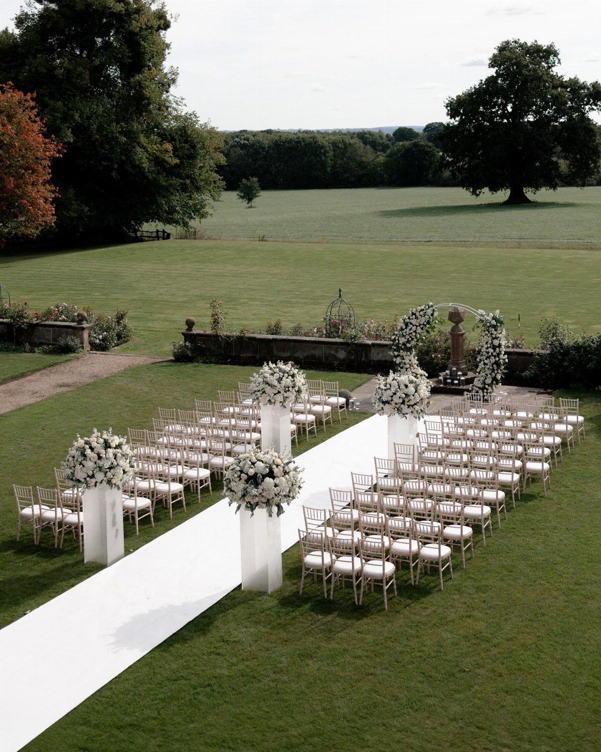 White wedding chairs and aisle runner arranged on a lush green lawn with floral arrangements.