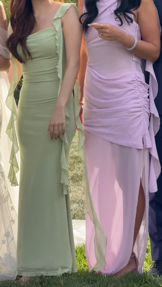 Two women standing in a garden wearing long, pastel-colored formal dresses with ruffled details.