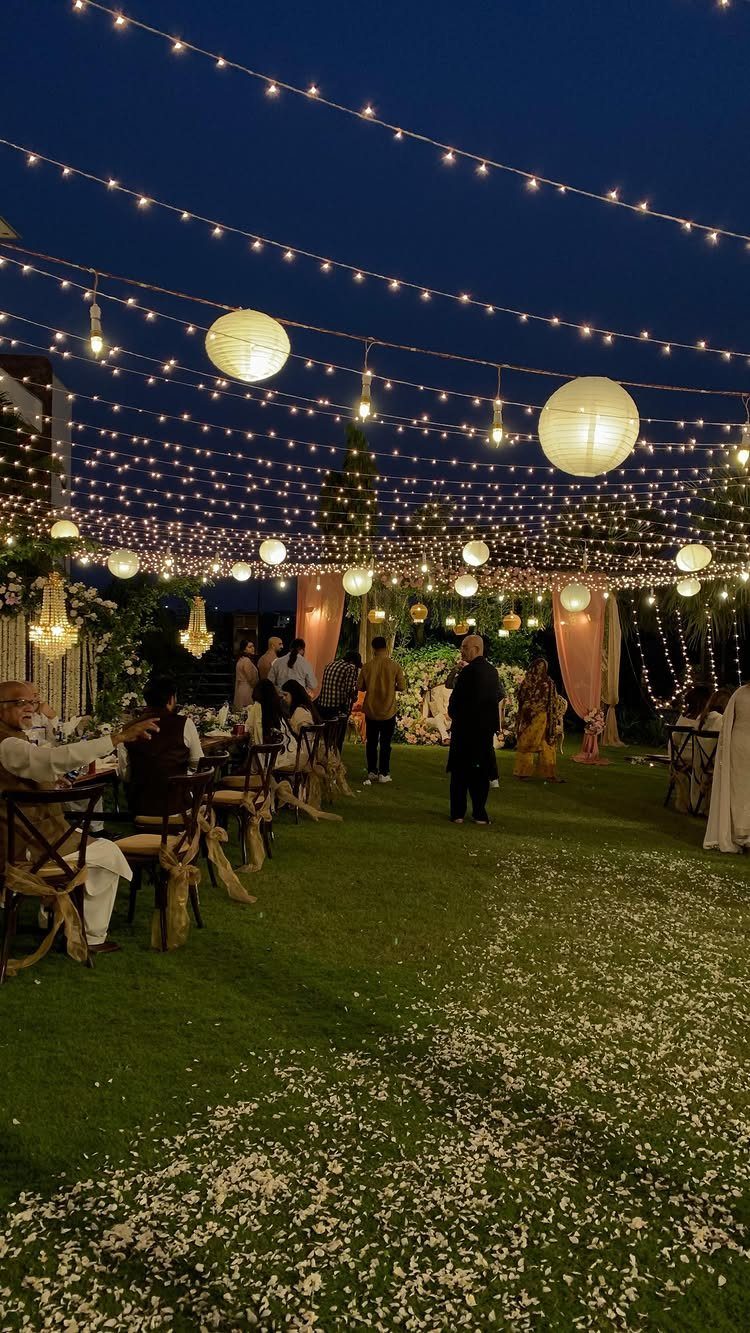Fairy lights and paper lanterns hang over guests seated at tables in a garden setting.