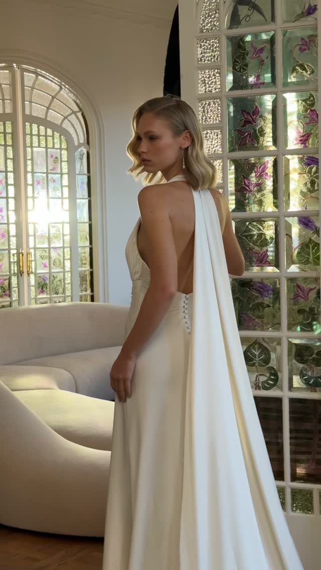 Glam Wedding Dress: 5 Looks for 2026 5 Blonde woman posing in a white backless halter wedding gown with a long flowing cape.