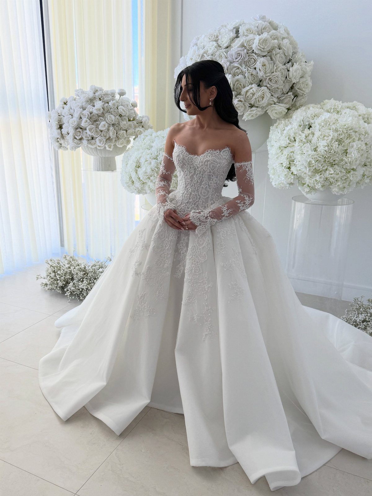 Glam Wedding Dress: 5 Looks for 2026 2 Woman in a white embellished off-the-shoulder ball gown standing among large white rose arrangements.