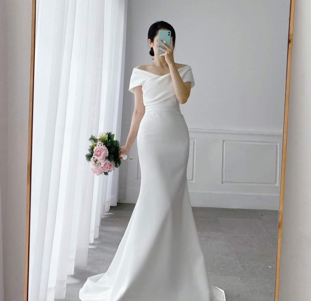 Glam Wedding Dress: 5 Looks for 2026 1 Woman in a white off-the-shoulder satin mermaid wedding gown holding pink roses.