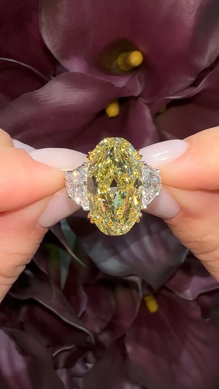 Gold and Champagne Wedding: 7 Ideas for a Warm, Metallic Celebration 2 Hands holding a large oval yellow diamond ring against dark purple flowers.
