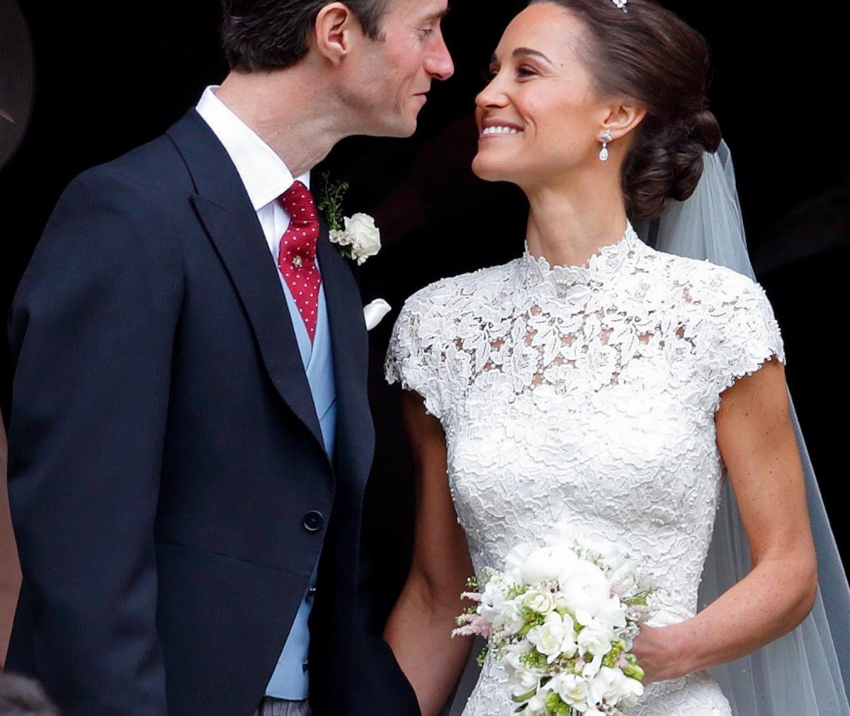 A smiling bride in a white lace cap sleeve gown holds a bouquet while looking at her groom.