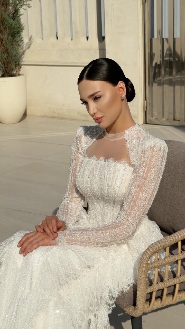 Woman sitting outdoors wearing a long-sleeved ivory lace wedding gown with a ruffled skirt.