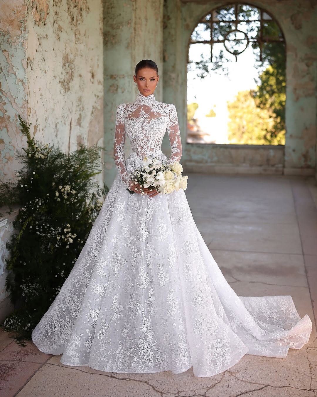A woman wearing a long-sleeved white lace wedding gown holding a white flower bouquet.