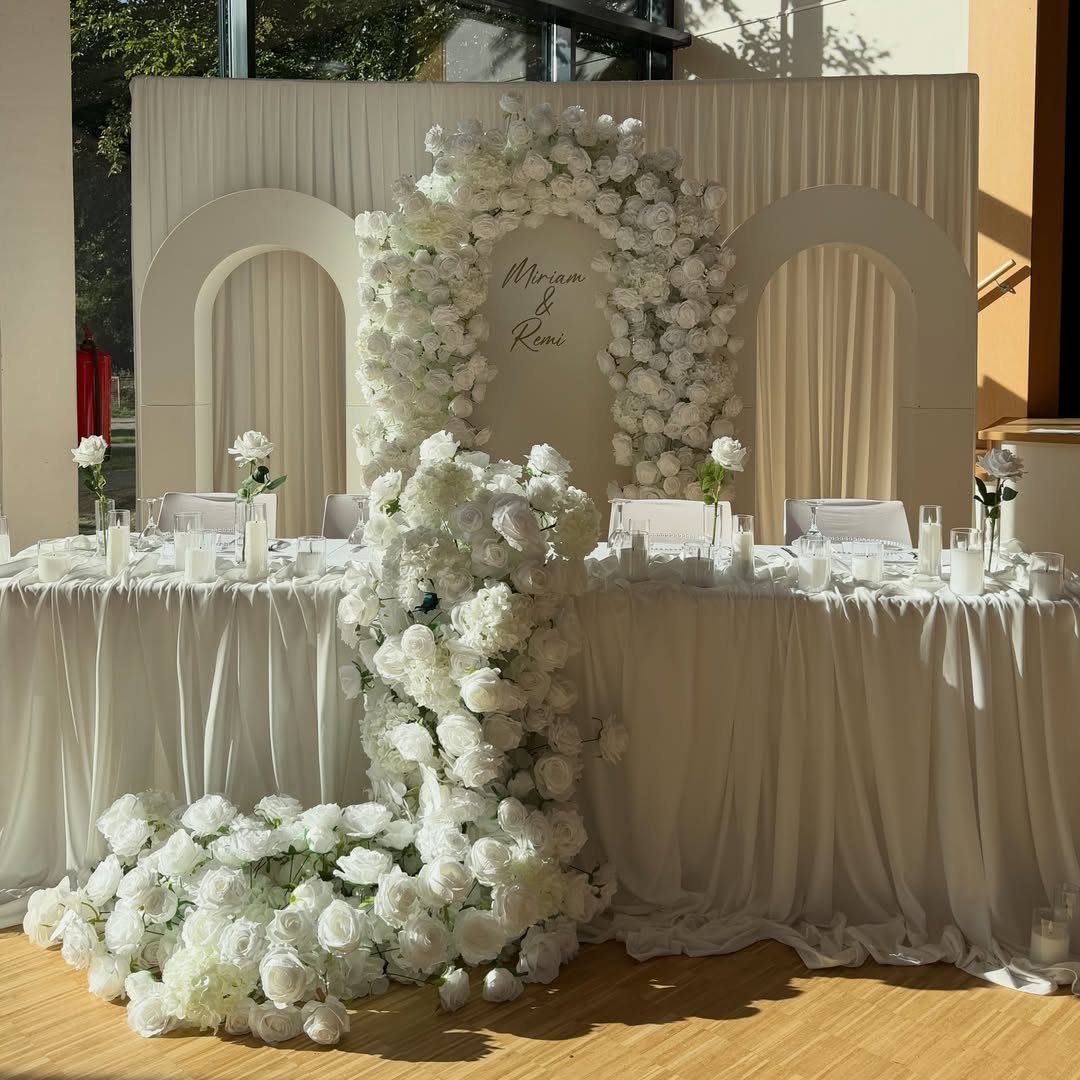 A long wedding head table decorated with white flowers, candles, and a large floral arch backdrop.