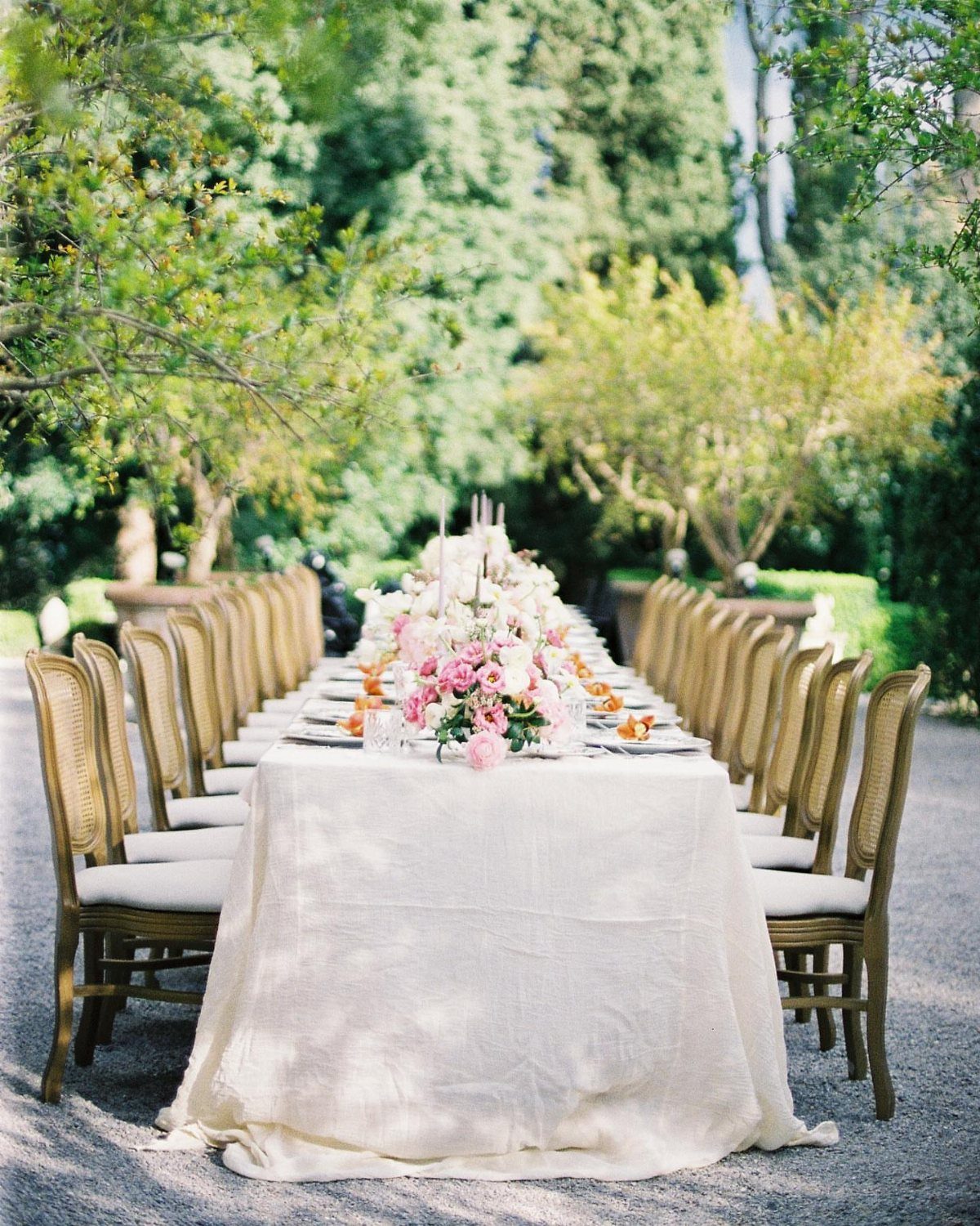 A long wedding table decorated with pink floral arrangements and white taper candles outdoors.