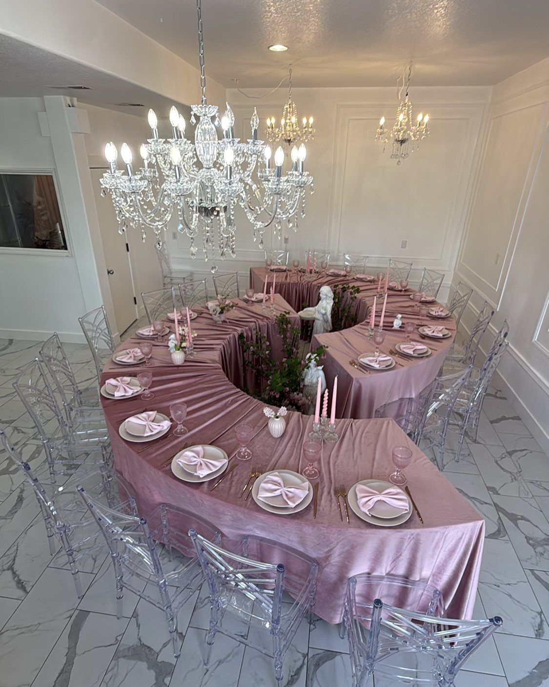 A curved serpentine wedding table set with rose satin linens, pink candles, and clear ghost chairs.