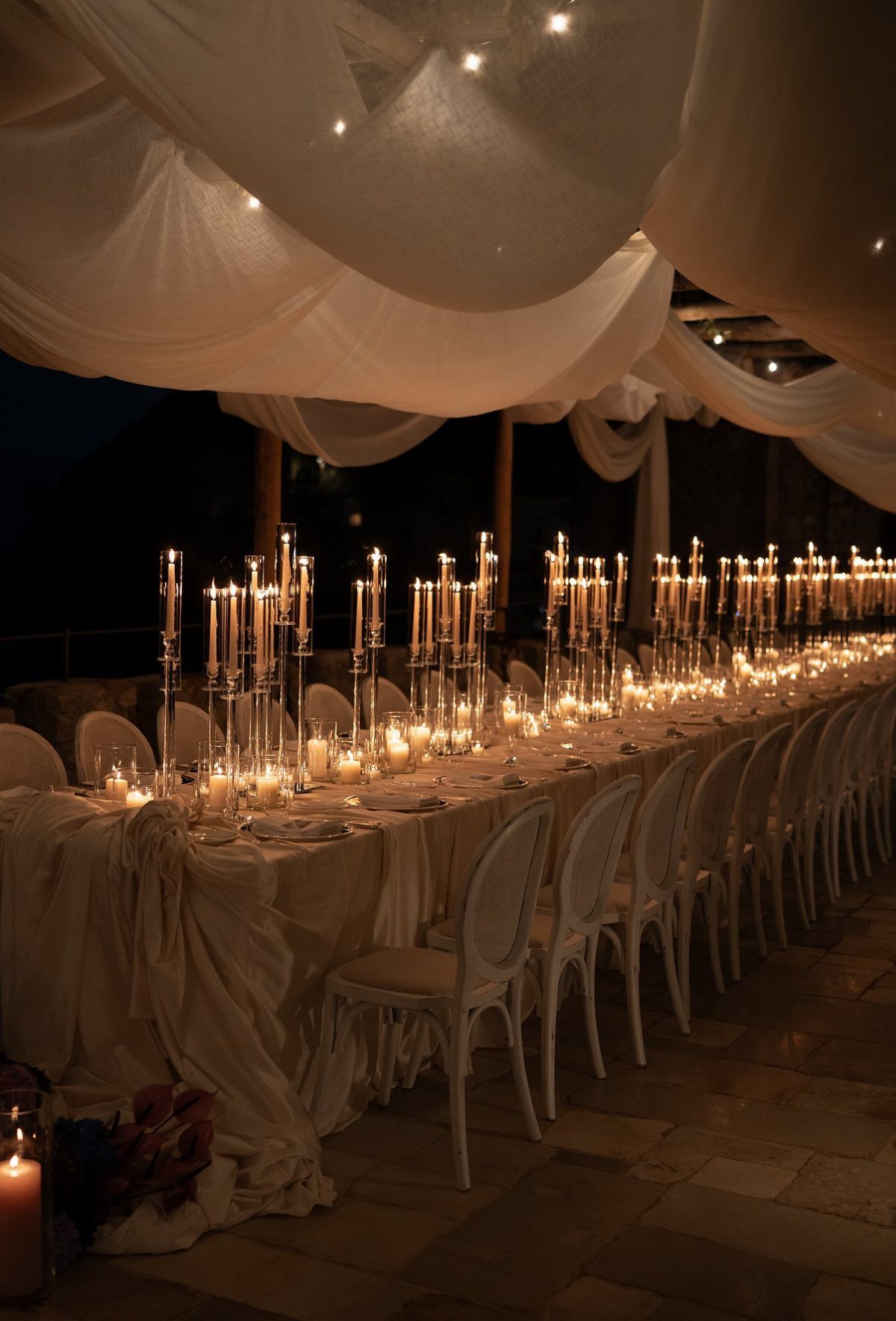 A long banquet table decorated with many tall white taper candles under white draped fabric.