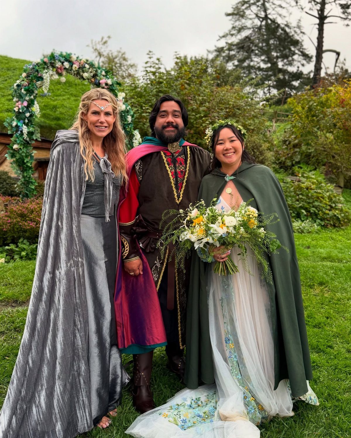Three people smiling in fantasy-themed wedding attire and capes in a lush green garden.