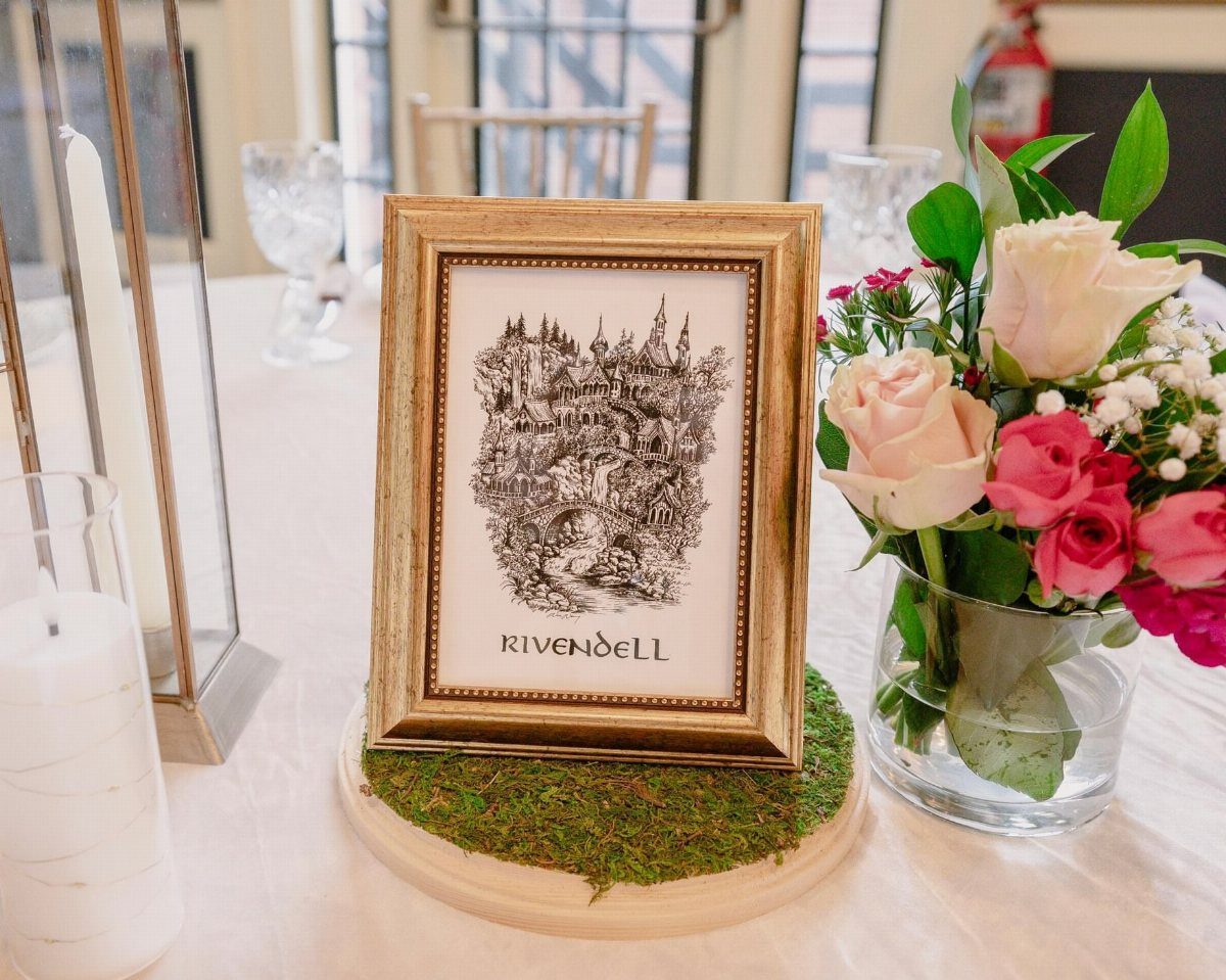 Framed Rivendell illustration on a mossy base next to pink roses on a wedding table.