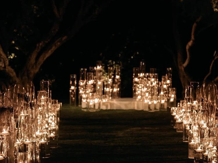 Glowing white candles in glass cylinders lining a dark outdoor path at night.
