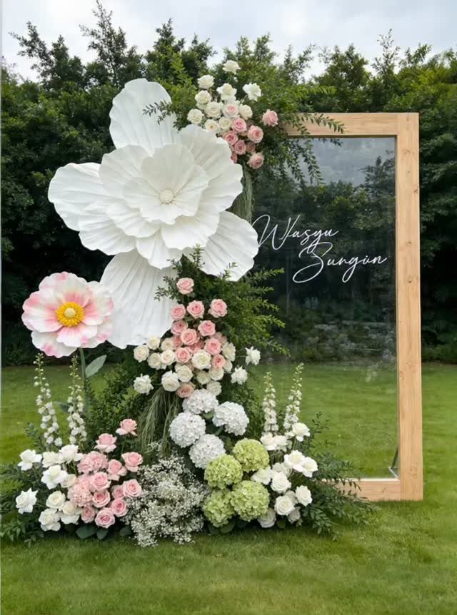 Large white paper flowers and pink floral arrangements decorating a wooden framed glass sign outdoors.