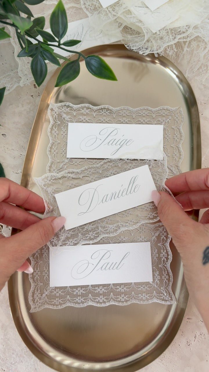 Hands holding elegant white place cards with cursive names over lace on a gold tray.