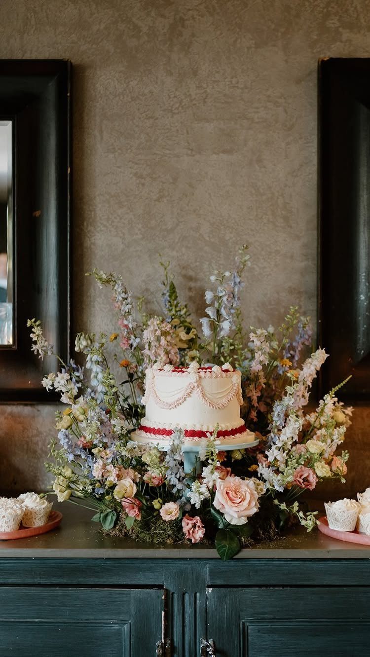 A white wedding cake decorated with pink beads, nestled within a large arrangement of pastel flowers.