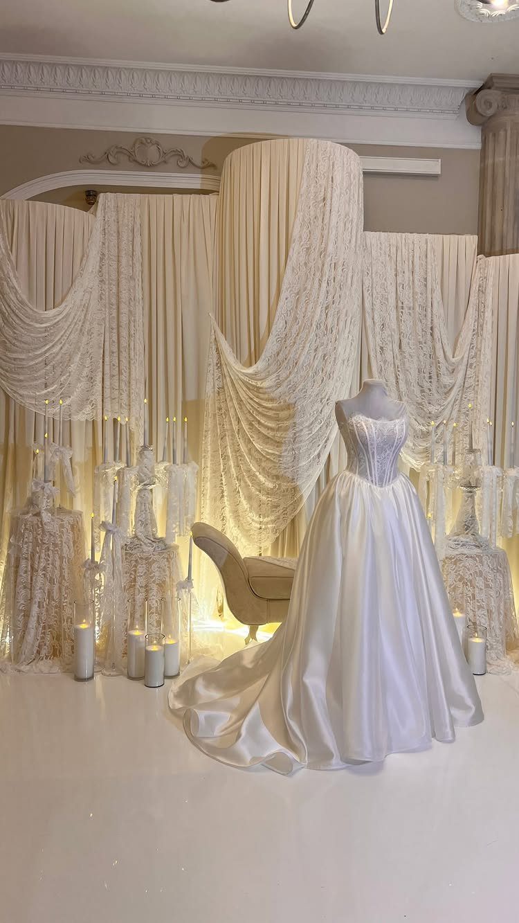A white wedding dress displayed in front of elegant, draped fabric backdrops and lit candles.