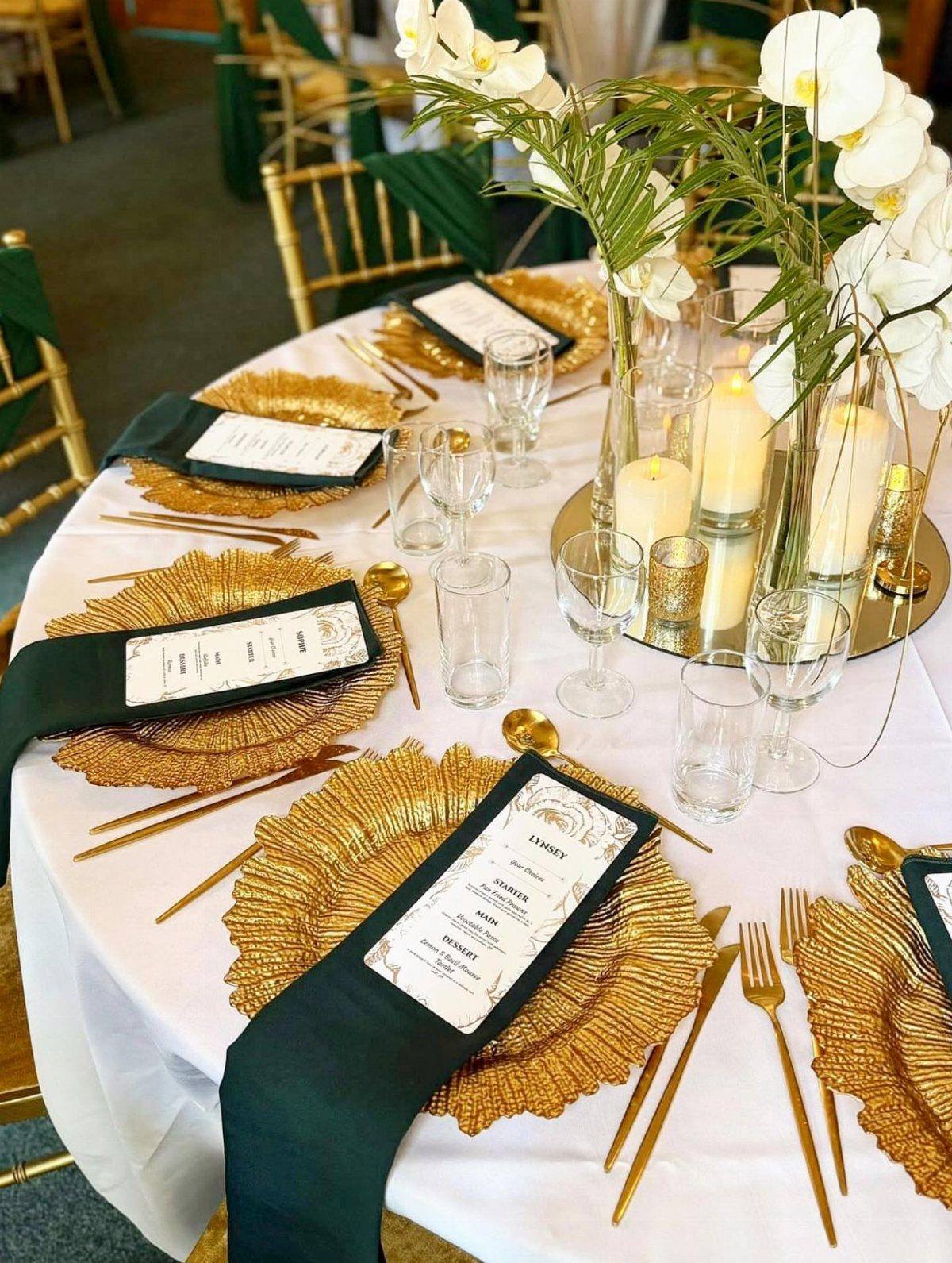 Elegant wedding table setting with gold chargers, green napkins, white orchids, and gold cutlery.