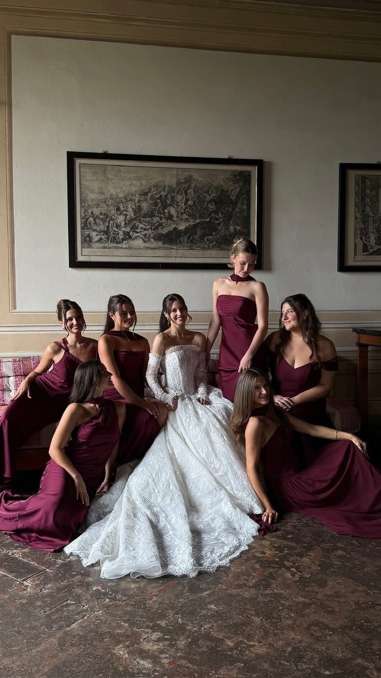 Bridesmaids in burgundy dresses pose around a bride wearing a white lace gown.