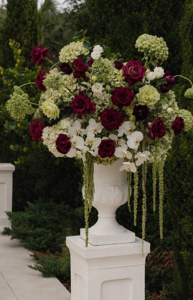 Burgundy roses and cream hydrangeas arranged in a white pedestal urn with trailing green vines.