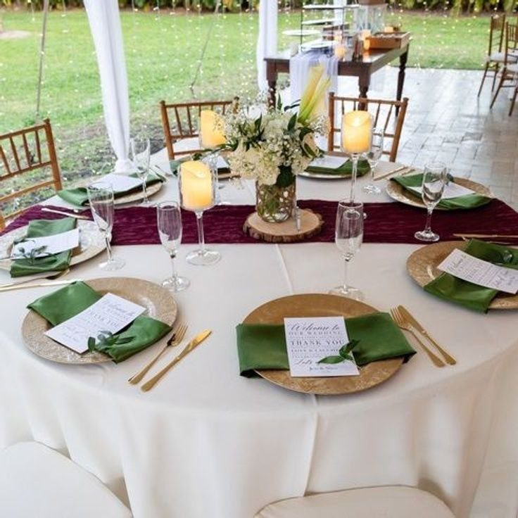 A wedding table set with gold chargers, green napkins, burgundy runner, and white floral centerpieces.