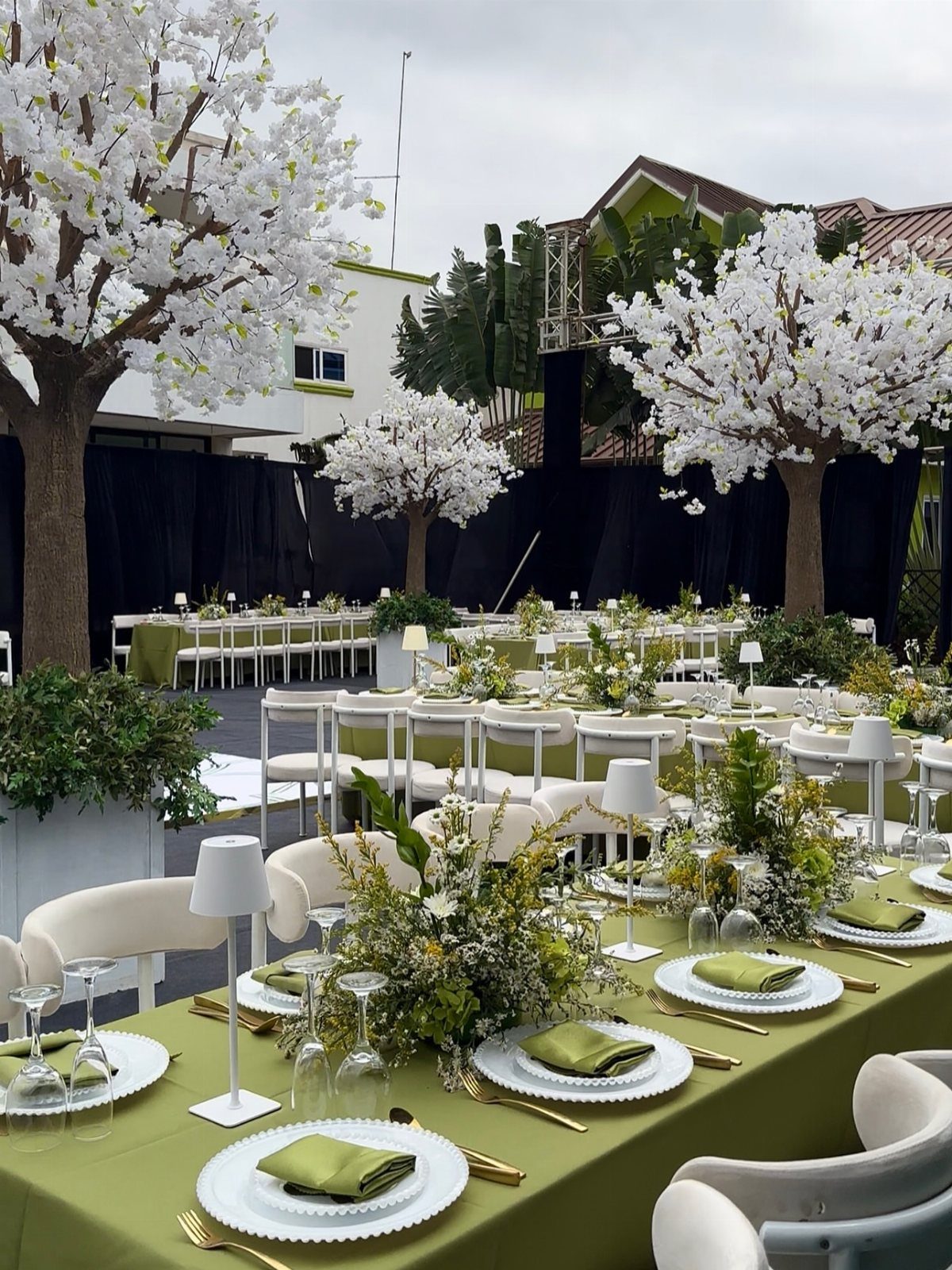Long dining tables covered in olive green linens with white floral centerpieces and gold cutlery outdoors.