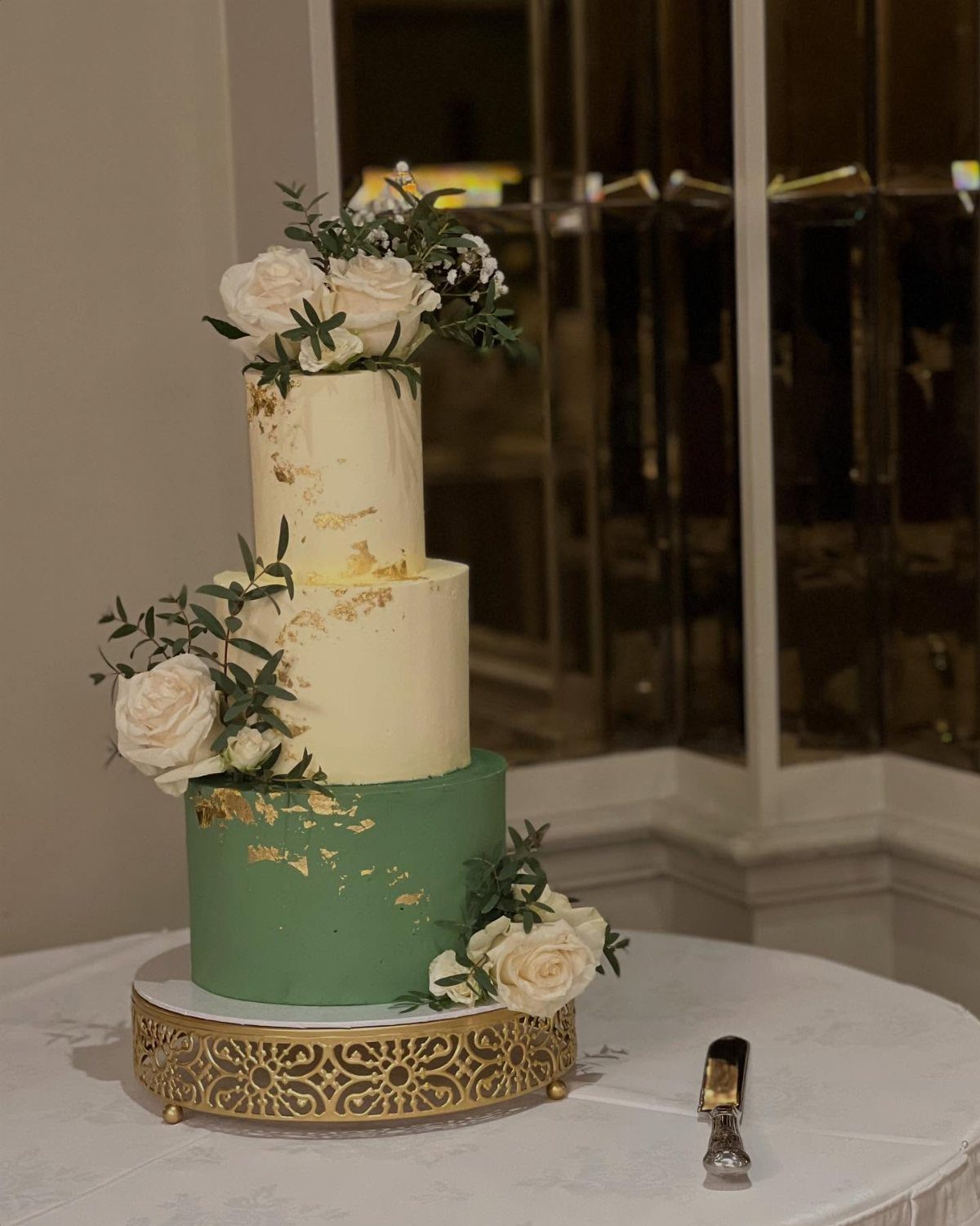 Three-tiered wedding cake with green and cream frosting, gold leaf, and white roses on a gold stand.
