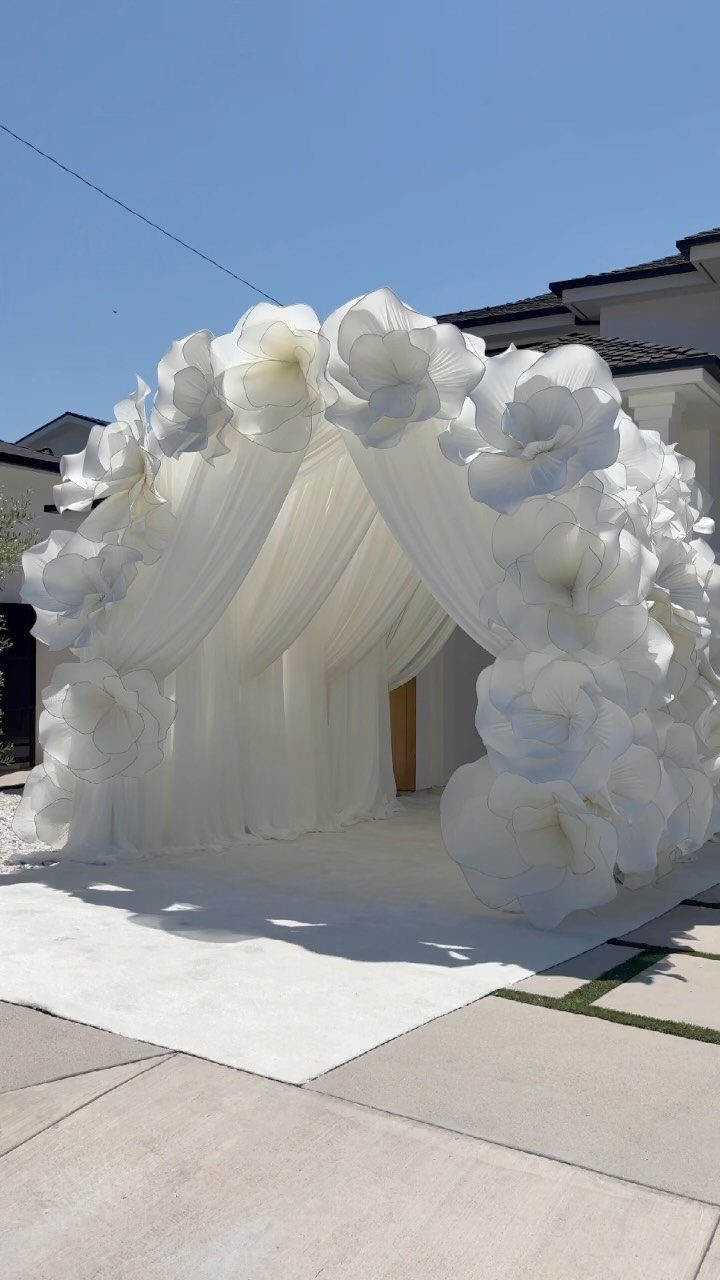 10 Outdoor Wedding Aisle Decor Ideas with Photos 7 Large white fabric floral arbor with flowing drapery standing on a white carpet outdoors.