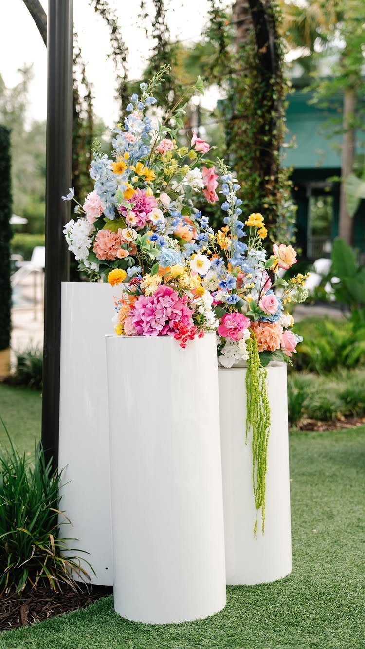 10 Outdoor Wedding Aisle Decor Ideas with Photos 6 Three white cylindrical pillars topped with vibrant, colorful floral arrangements on a green lawn.