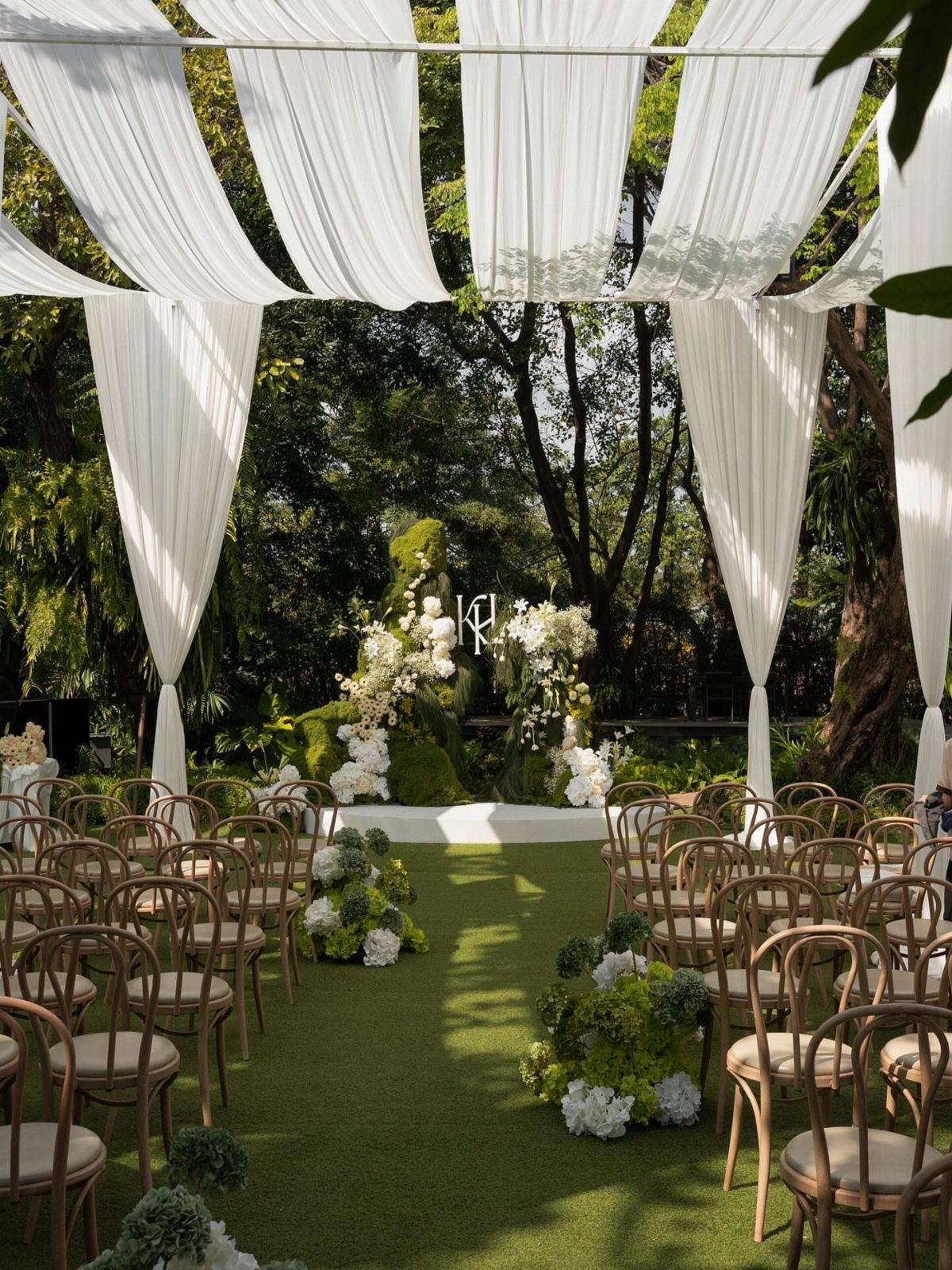 10 Outdoor Wedding Aisle Decor Ideas with Photos 1 White fabric drapes hang over an outdoor wedding ceremony aisle lined with white flowers and wooden chairs.