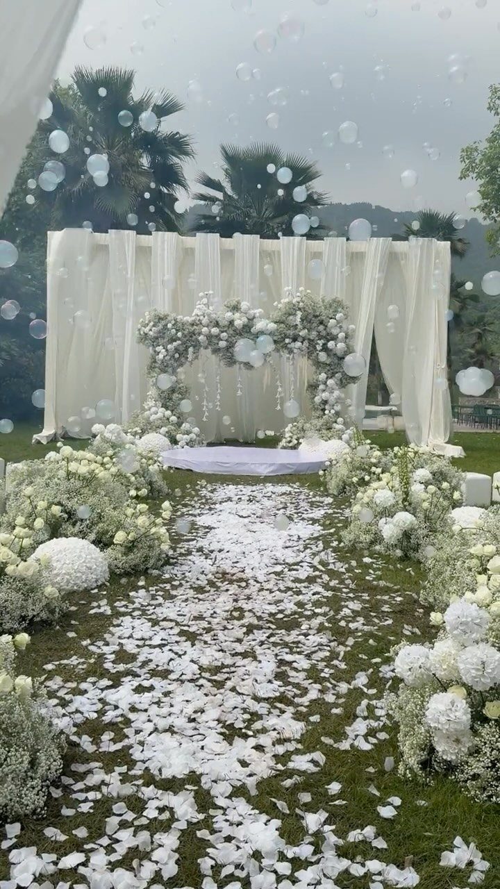 White flower petals line a grass aisle leading toward a white fabric wedding backdrop with floral garlands.