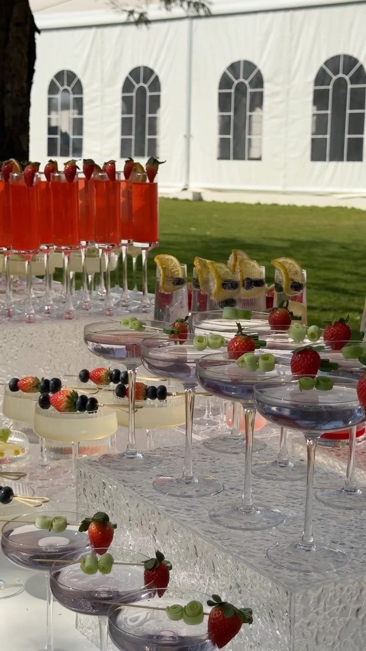 Cocktail glasses with red and clear drinks garnished with strawberries and grapes on a white table.