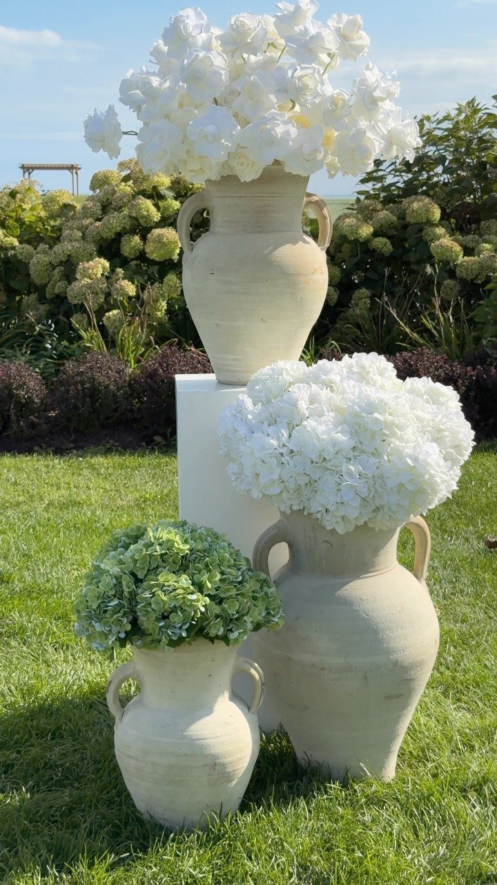 Three large ceramic vases filled with white and green hydrangea floral arrangements on a green lawn.