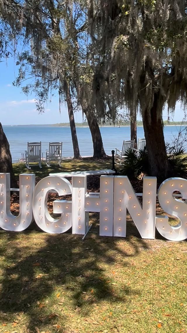 Large white marquee letters sit on green grass near a waterfront with trees.